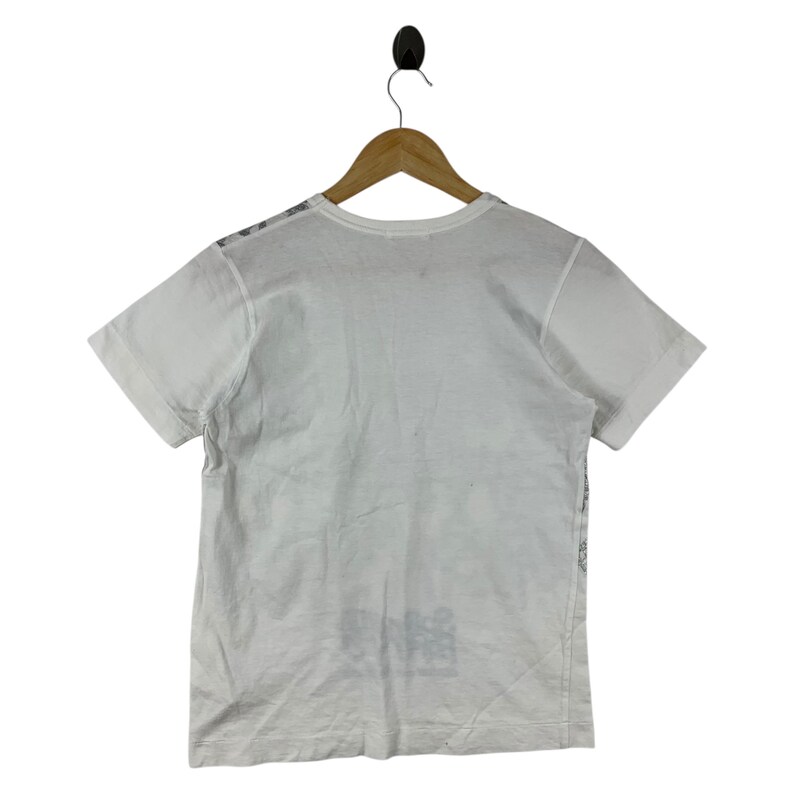 Puede incluir: Camiseta blanca de manga corta colgada de una percha de madera. La camiseta tiene cuello redondo y un dise&ntilde;o gr&aacute;fico en la parte inferior delantera. La camiseta est&aacute; hecha de un material suave y ligero.