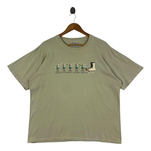 Peut inclure: Un t-shirt vert clair avec un imprimé graphique d'un groupe de personnages en bâtonnets portant des costumes verts et bleus, soulevant des poids, et un pied sur des patins à roulettes. Le texte "MONTY PYTHON'S FLYING CIRCUS" est imprimé sous le graphique.