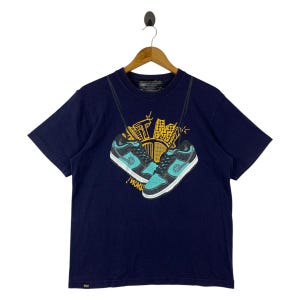 May include: A navy blue t-shirt with a graphic of two trainers, teal and black in colour, hanging in front of a yellow and gold cityscape. The text "!WORK" is visible below the trainers.