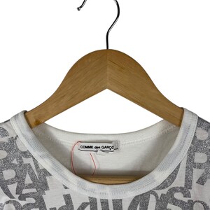 Puede incluir: Camiseta blanca con cuello redondo y la inscripci&oacute;n "COMME des GAR&Ccedil;ONS" en la etiqueta. La camiseta presenta un patr&oacute;n de letras plateadas con purpurina. La prenda est&aacute; colgada de una percha de madera.