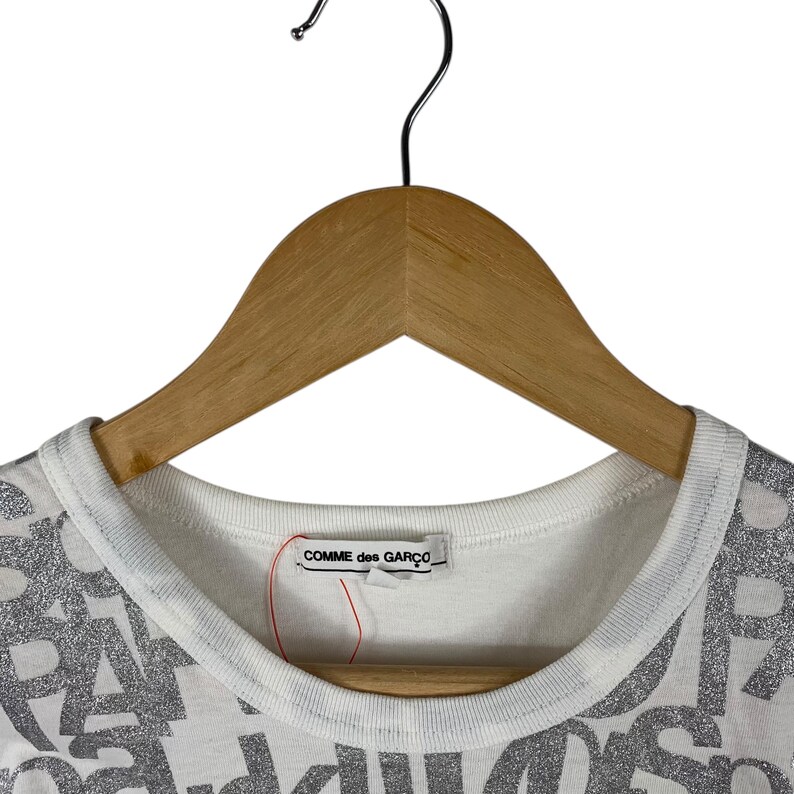 Puede incluir: Camiseta blanca con cuello redondo y la inscripci&oacute;n "COMME des GAR&Ccedil;ONS" en la etiqueta. La camiseta presenta un patr&oacute;n de letras plateadas con purpurina. La prenda est&aacute; colgada de una percha de madera.