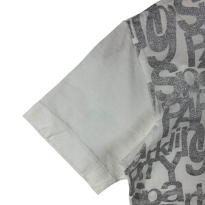 Puede incluir: Primer plano de una manga de camiseta blanca. El lado derecho de la camiseta presenta un dise&ntilde;o plateado con purpurina con la palabra "party" visible. La manga es de color blanco liso.