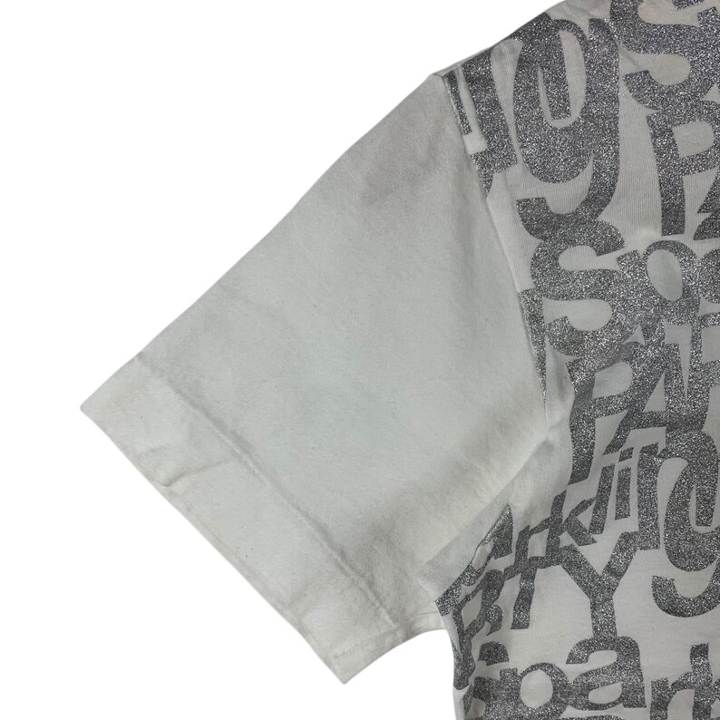 Puede incluir: Primer plano de una manga de camiseta blanca. El lado derecho de la camiseta presenta un dise&ntilde;o plateado con purpurina con la palabra "party" visible. La manga es de color blanco liso.