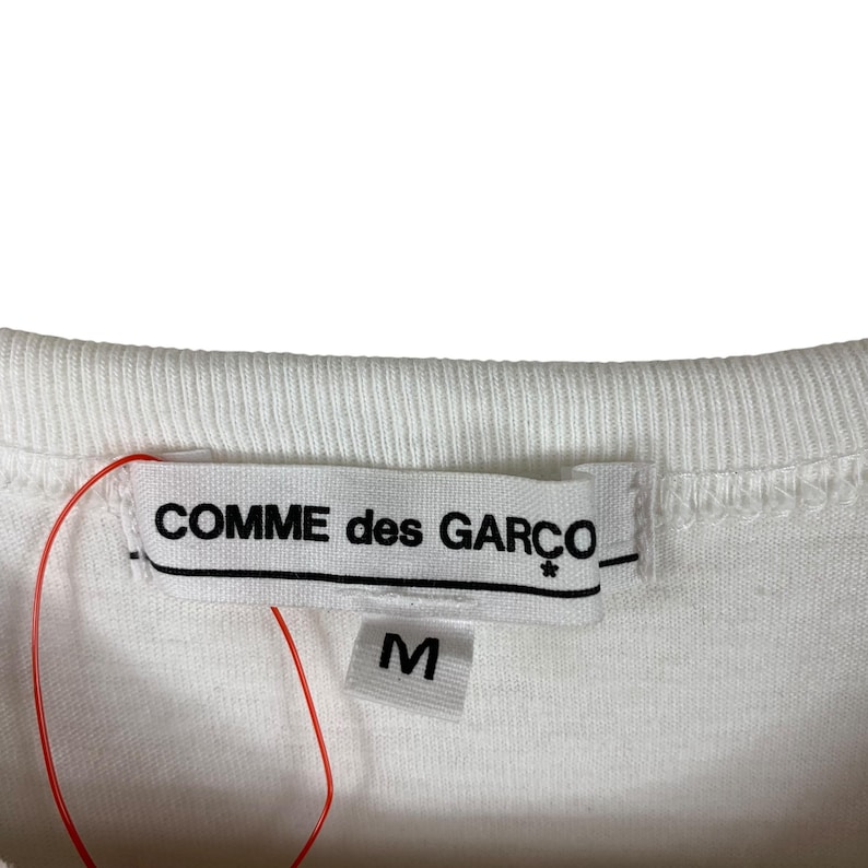 Puede incluir: Primer plano de un cuello de camiseta blanca con una etiqueta que dice "COMME des GAR&Ccedil;ONS" en texto negro. Debajo de la etiqueta hay una etiqueta m&aacute;s peque&ntilde;a con la letra "M". Una cuerda roja est&aacute; unida a las etiquetas.