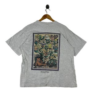 May include: A light grey t-shirt with a vibrant graphic print on the back. The design showcases cartoon characters, plants, and the text "OCTOPUS ARMY WILDLIFE" and "Delicious Army". The t-shirt is suspended from a wooden hanger.