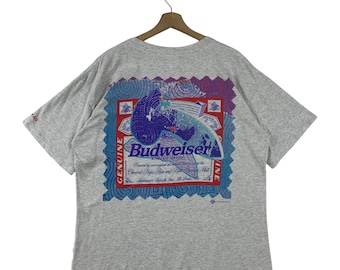 May include: A heather gray t-shirt featuring a large graphic on the back. The graphic is a stylized Budweiser label with blue, red, and white elements. The text "Budweiser" is prominently displayed.