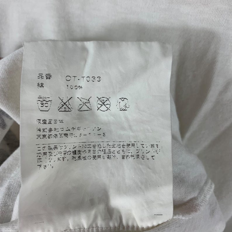 Puede incluir: Primer plano de una etiqueta de tela blanca con texto japon&eacute;s y s&iacute;mbolos de cuidado. La etiqueta incluye el c&oacute;digo de producto "OT-T033" y la composici&oacute;n del material "100%" algod&oacute;n. Instrucciones de lavado visibles.