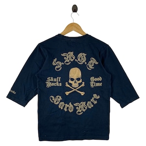 May include: Navy blue long-sleeve shirt with a gold skull and crossbones graphic on the back. The shirt has the text "Skull Works Good Time Hard Ware" in gold lettering. The sleeves are three-quarter length.