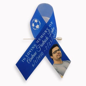 Peut inclure: Un ruban de sensibilisation bleu avec un motif de ballon de football blanc, le texte IN LOVING MEMORY OF Andrew Fredrick Lake 6/1/1950 - 7/23/2023, et une photo d'une personne portant des lunettes, fix&eacute; par une &eacute;pingle en m&eacute;tal argent&eacute;.