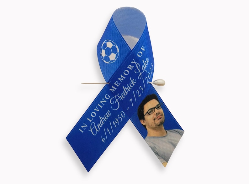 Peut inclure: Un ruban de sensibilisation bleu avec un motif de ballon de football blanc, le texte IN LOVING MEMORY OF Andrew Fredrick Lake 6/1/1950 - 7/23/2023, et une photo d'une personne portant des lunettes, fix&eacute; par une &eacute;pingle en m&eacute;tal argent&eacute;.