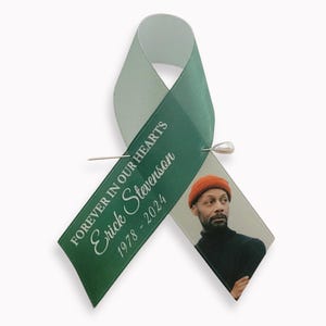 Peut inclure: Un ruban de sensibilisation vert fonc&eacute; avec une &eacute;pingle argent&eacute;e. Le ruban porte le texte blanc "FOREVER IN OUR HEARTS", le nom "Erick Stevenson" et les ann&eacute;es "1978 - 2024". Une photographie d'une personne portant un bonnet orange est fix&eacute;e.