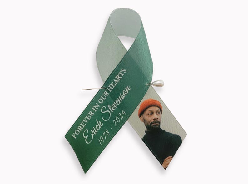 Peut inclure: Un ruban de sensibilisation vert fonc&eacute; avec une &eacute;pingle argent&eacute;e. Le ruban porte le texte blanc "FOREVER IN OUR HEARTS", le nom "Erick Stevenson" et les ann&eacute;es "1978 - 2024". Une photographie d'une personne portant un bonnet orange est fix&eacute;e.