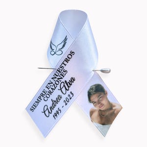 Peut inclure: Un ruban de sensibilisation en satin blanc orn&eacute; d'un motif noir repr&eacute;sentant des ailes d'ange au-dessus d'un symbole infini. Le texte indique SIEMPRE EN NUESTROS CORAZONES, Andrea Alva, 1995-2023. Une photo est imprim&eacute;e sur le pan droit, fix&eacute; par une &eacute;pingle.