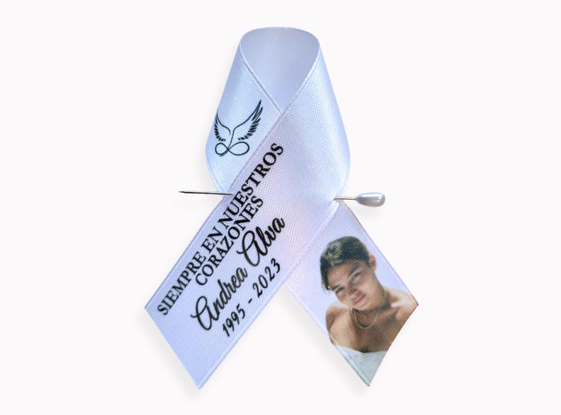 Peut inclure: Un ruban de sensibilisation en satin blanc orn&eacute; d'un motif noir repr&eacute;sentant des ailes d'ange au-dessus d'un symbole infini. Le texte indique SIEMPRE EN NUESTROS CORAZONES, Andrea Alva, 1995-2023. Une photo est imprim&eacute;e sur le pan droit, fix&eacute; par une &eacute;pingle.