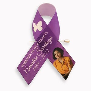 Peut inclure: Un ruban de sensibilisation violet orn&eacute; d'un papillon blanc, du texte "FOREVER IN OUR HEARTS", du nom "Annalise Saratoga" et des ann&eacute;es "1989 - 2022". Une photo d'une personne en chemise orange est imprim&eacute;e sur le ruban, fix&eacute; par une &eacute;pingle.