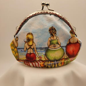 May include: A coin purse with a silver clasp and a colorful beach scene. The fabric features four figures in swimsuits sitting on a beach, with a blue sky and ocean in the background. The purse is on a white surface.