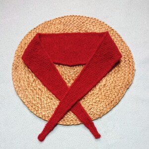 Peut inclure: Une écharpe rouge vif tricotée, présentée sur un tapis rond et tissé de couleur beige. Le design de l'écharpe présente une section large et rectangulaire qui se rétrécit en deux longues extrémités pointues, se croisant. Le fond est une surface bleu clair.