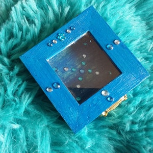 Puede incluir: Una pequeña caja de madera azul con un interior de espejo. La caja está decorada con diamantes de imitación y tiene un cierre dorado.