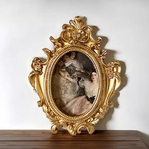 May include: An ornate gold picture frame with an oval aperture. The frame has detailed floral and scrollwork patterns. The framed artwork shows a classical painting of three women in elegant clothing. The frame is displayed on a wooden surface.