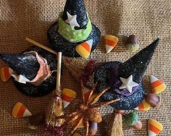 May include: A collection of Halloween decorations including three black glittered witch hats with star accents, candy corn, acorns, a glittery spider, and miniature brooms. The hats have green and purple bands.