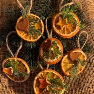 Puede incluir: Seis adornos navideños hechos a mano con rodajas de naranja secas, decorados con vegetación y pequeñas formas de fieltro. Cada adorno tiene un lazo de cuerda para colgar. Los adornos están dispuestos sobre agujas de pino y arpillera.