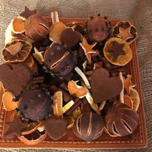 May include: A square, orange ceramic dish filled with dried orange slices, star-shaped spices, heart-shaped decorations, and cinnamon sticks. The arrangement is a mix of brown, orange, and tan colors, creating a rustic, aromatic display.