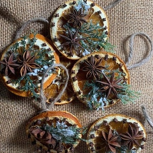 May include: Dried orange slice decorations with star anise, cloves, and greenery. Each decoration has a twine loop for hanging. The decorations are arranged on a burlap surface, creating a rustic, natural aesthetic.