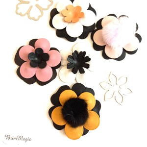 May include: Five felt flower embellishments in various colors, including black, white, pink, orange, and yellow. Each flower has a center pom-pom in a contrasting color. The flowers are arranged on a white background.