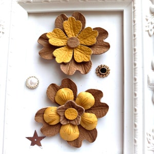 Puede incluir: Dos flores de cuero marrón con centros amarillos. Las flores están superpuestas y tienen una superficie texturizada. Las flores se muestran sobre un fondo blanco.