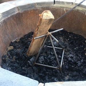 May include: A black metal fire grate with a wooden log resting on top. The grate is sitting in a round fire pit filled with charcoal.