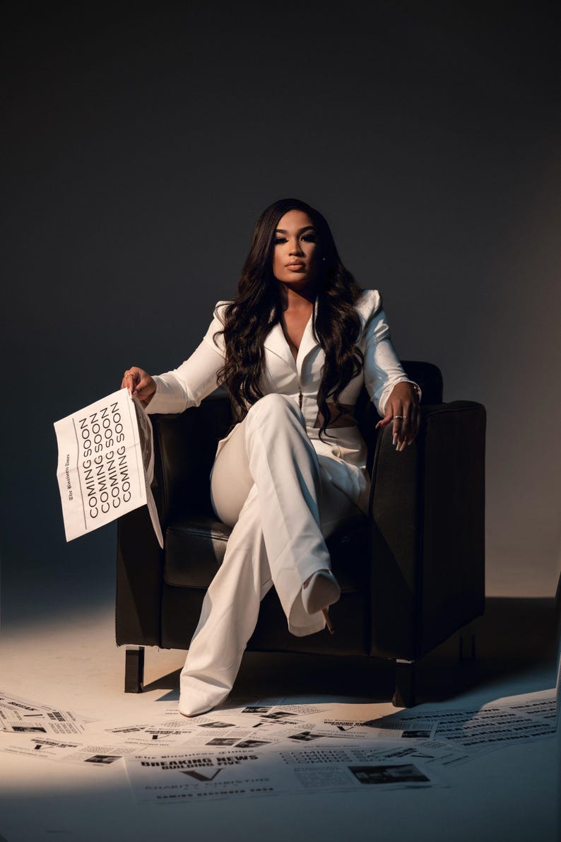 May include: A woman in a white pantsuit sits in a black leather chair, holding a newspaper with the text "Coming Soon" repeated multiple times. The newspaper is surrounded by other newspapers on the floor.