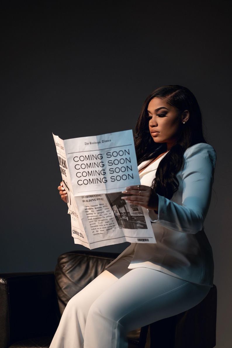 May include: A woman in a white blazer and pants is sitting on a black leather chair. She is reading a newspaper with the headline "Coming Soon" repeated four times. The newspaper also has a photo of a building with the text "Introducing Building Five!"