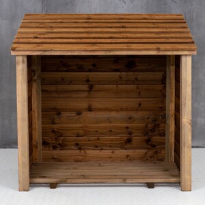May include: A wooden log storage shed with a slanted roof and a slatted design. The shed is empty and has a natural wood finish.