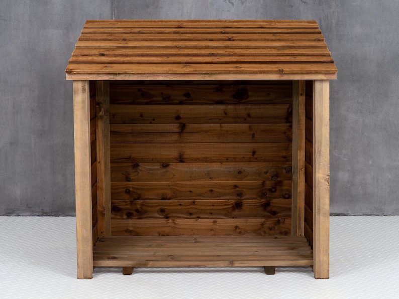 May include: A wooden log storage shed with a slanted roof and a slatted design. The shed is empty and has a natural wood finish.