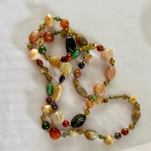 May include: A long necklace made of various colored stones, including red, orange, yellow, green, brown, and purple. The stones are all different shapes and sizes, and they are strung together on a gold chain.