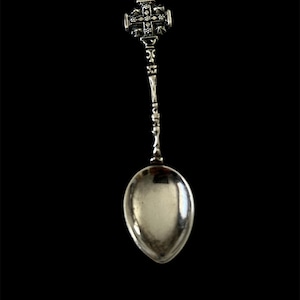 Peut inclure: Une cuillère en argent avec un motif de croix et de couronne décoratif sur le manche. La cuillère a un bol ovale et un long manche orné. La cuillère est probablement en argent sterling ou en métal similaire.