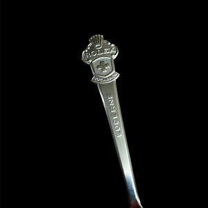 May include: A silver spoon with the word "LUCERNE" engraved on the handle. The spoon's head features the Rolex logo and a shield with a cross, with the word "BUCHERER" below the shield. The spoon is set against a black background.
