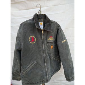 May include: A dark gray Carhartt jacket with a full zipper closure and a collar. The jacket features embroidered text and logos, including "National High School Rodeo Association" and "Event Champion". A small Carhartt logo is on the sleeve.