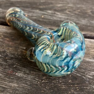May include: A hand-blown glass pipe with a swirling pattern of teal, blue, and gold. The pipe features a rounded bowl and a curved stem, with a small glass handle on the side. The glass has a glossy finish.