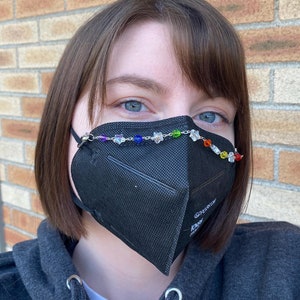 May include: A person wearing a black face mask with a rainbow beaded chain across the top. The person is wearing a dark grey hoodie.