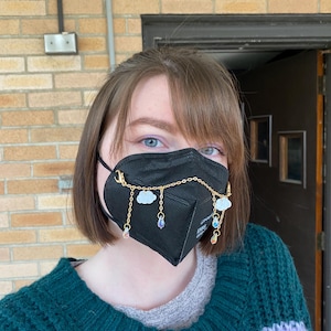 Op de afbeelding: Een persoon draagt ​​een zwart mondkapje met een gouden ketting en bedels. De bedels zijn in de vorm van wolken en regendruppels. De persoon draagt ​​een groen en paarse trui.