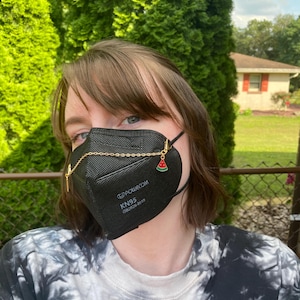 May include: Black KN95 face mask with a gold chain and watermelon charm. The mask has the text "POLWECOM KN95 GB2626-2019". The person is wearing a tie-dye shirt. Green foliage is in the background.