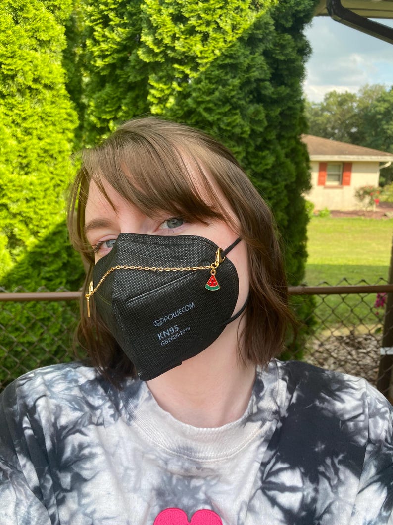 May include: Black KN95 face mask with a gold chain and watermelon charm. The mask has the text "POLWECOM KN95 GB2626-2019". The person is wearing a tie-dye shirt. Green foliage is in the background.