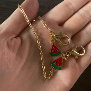 May include: Gold-colored chain with two lobster clasps and two watermelon slice charms. The watermelon charms are red with black seeds and green rinds. The chain is held in a hand.