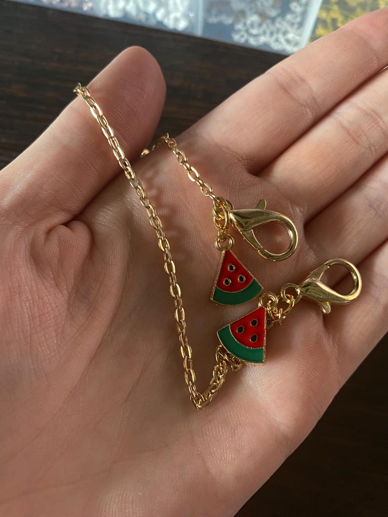 May include: Gold-colored chain with two lobster clasps and two watermelon slice charms. The watermelon charms are red with black seeds and green rinds. The chain is held in a hand.