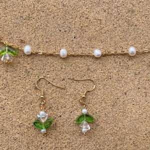 May include: A gold chain necklace with white pearl beads and green flower charms. Matching earrings with green flower charms and white pearl beads.