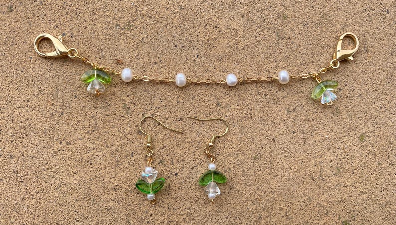 May include: A gold chain necklace with white pearl beads and green flower charms. Matching earrings with green flower charms and white pearl beads.