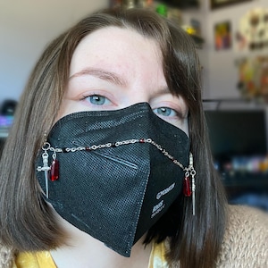 May include: A person wearing a black face mask with a silver chain and red and silver charms attached. The chain is draped over the mask and the charms hang down on either side. The mask has the text "KN95" and "Designed in Korea" printed on it.