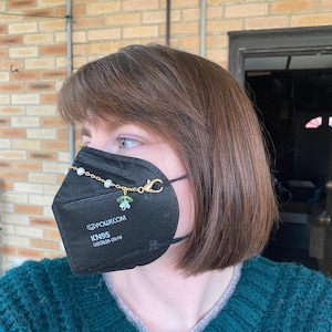May include: A black face mask with a gold chain and green and white beads attached to the ear loops. The mask has the text "POWECOM KN95 GB2626-2019" printed on it.