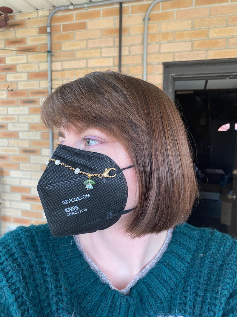 May include: A black face mask with a gold chain and green and white beads attached to the ear loops. The mask has the text "POWECOM KN95 GB2626-2019" printed on it.